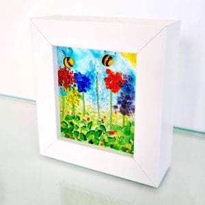 A small white box frame featuring a wildflower meadow scene with handmade glass bees.