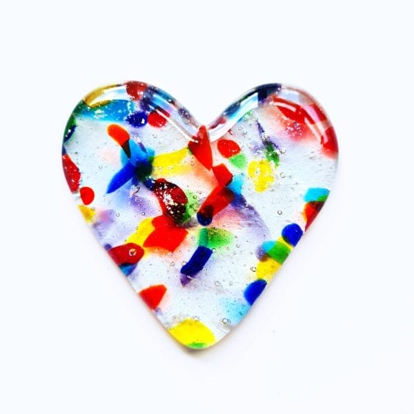 Cremation Ashes fused inside a clear glass heart with small pieces of coloured glass, on a white background.