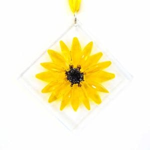 A yellow sunflower made of glass on a clear glass base with cremation ashes fused inside the glass, hanging with yellow ribbon on a white wall.