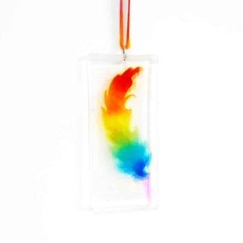 A Rainbow coloured feather on clear glass with rainbow ribbon, hanging on a white background.