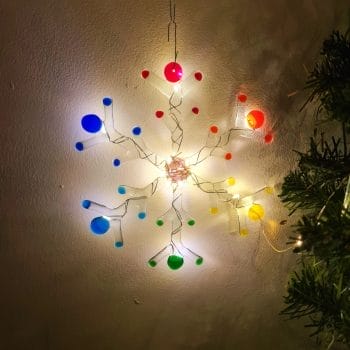 A handmade fused glass snowflake with rainbow colours and warm white leds on a white background with a green christmas tree.