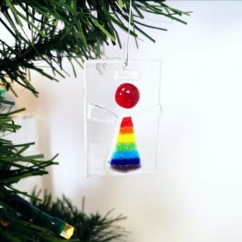 A fused glass Angel christmas ornament with a rainbow robe and red head, with oridescent wings hanging from a green christmas tree branch.