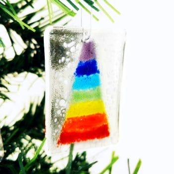 A fused glass hanging christmas decoration featuring a rainbow christmas tree and white "snow" hanging on a white background hanging from a green christmas tree.